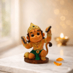 Smiling Bal Hanuman Ji seated figurine in vibrant multicolor finish with golden crown and ornaments, holding cymbals, placed on marble surface with diya in background